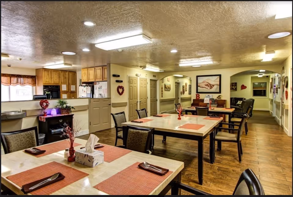 Interior view of a dining area in a senior living facility with several tables set with orange placemats, napkins, and utensils. The room has wooden flooring, beige walls, and ceiling lights. In the background, there is a kitchen area with wooden cabinets and a refrigerator. The walls are decorated with framed pictures and heart-shaped decorations.