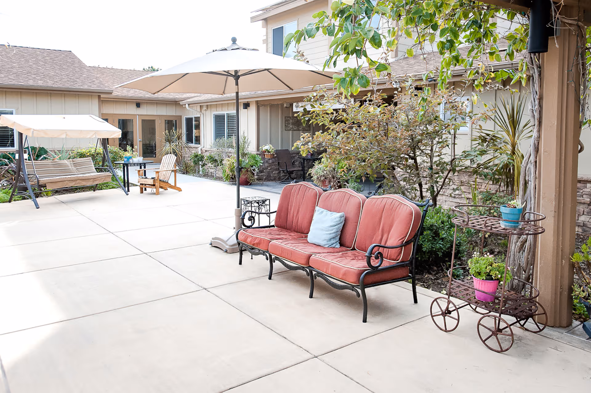 Sunlit outdoor courtyard with a red-cushioned bench, umbrella, swing, potted plants, and patio seating in front of a senior living building.