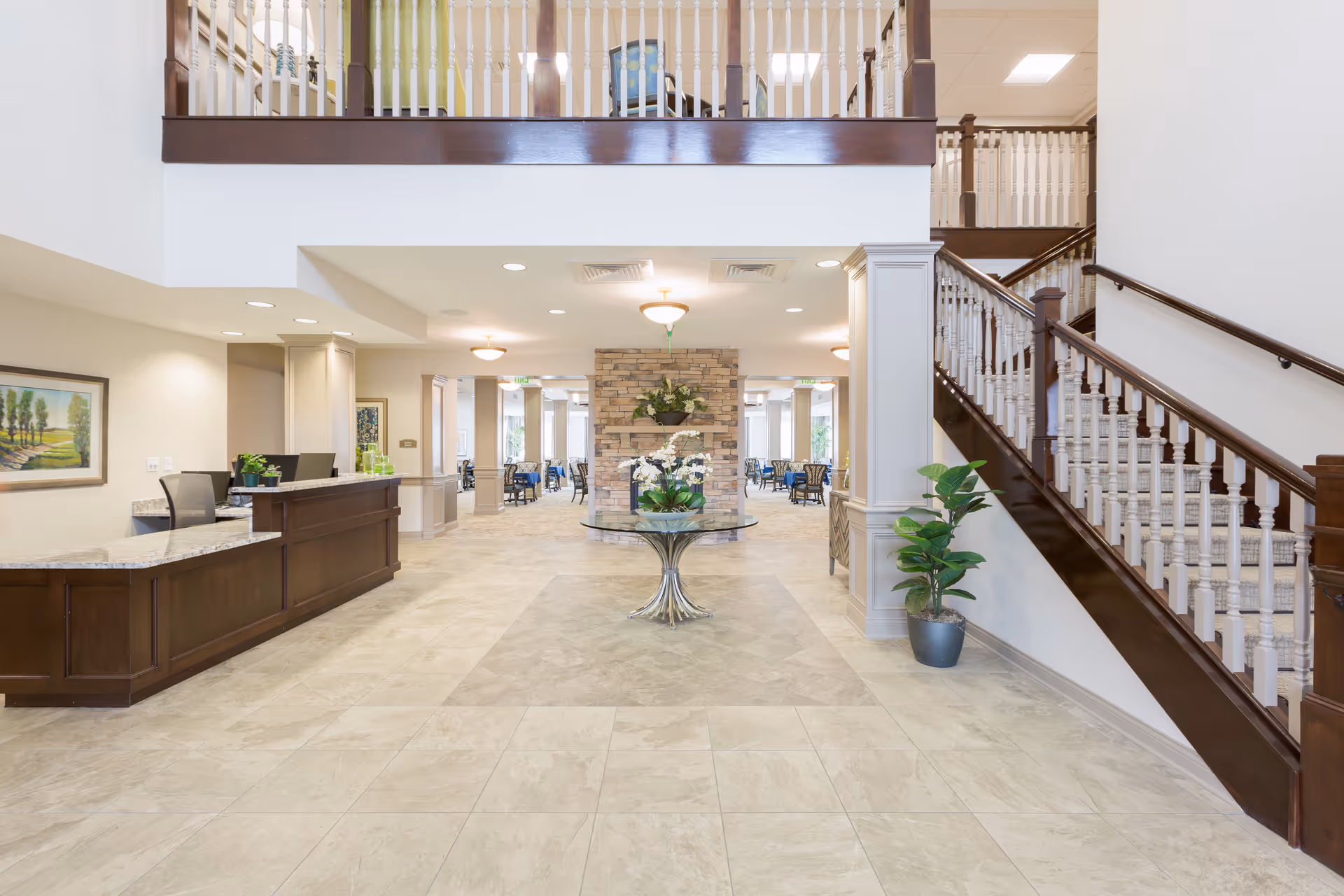 Bright senior living lobby with a reception desk on the left, a central table with flowers, and a staircase on the right leading to an upper balcony.