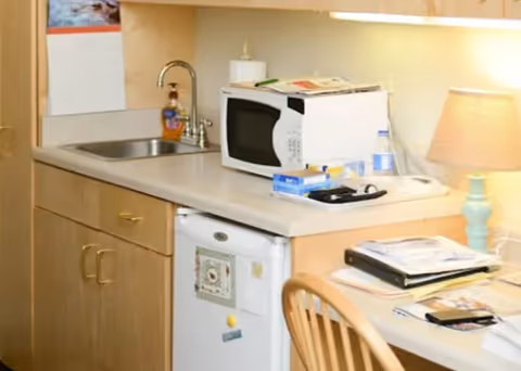 A small kitchenette area with a sink, microwave, mini refrigerator, and countertop. There are various items on the counter including a lamp, water bottle, soap dispenser, and some papers. A wooden chair is partially visible in front of the counter.