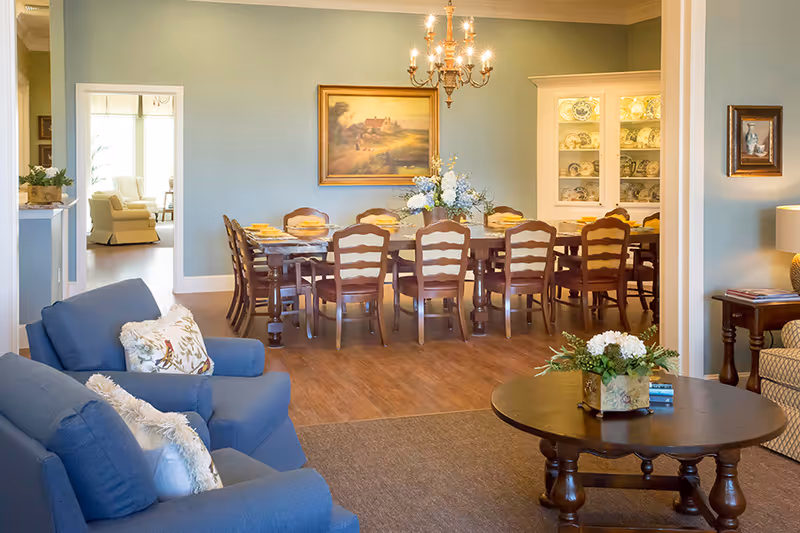 A cozy interior view showing a living room with blue armchairs and a round wooden coffee table with a floral arrangement. Beyond the living room is a dining room with a long wooden table set with yellow plates and surrounded by wooden chairs. The dining room features a chandelier, a painting on the wall, and a cabinet displaying decorative plates.