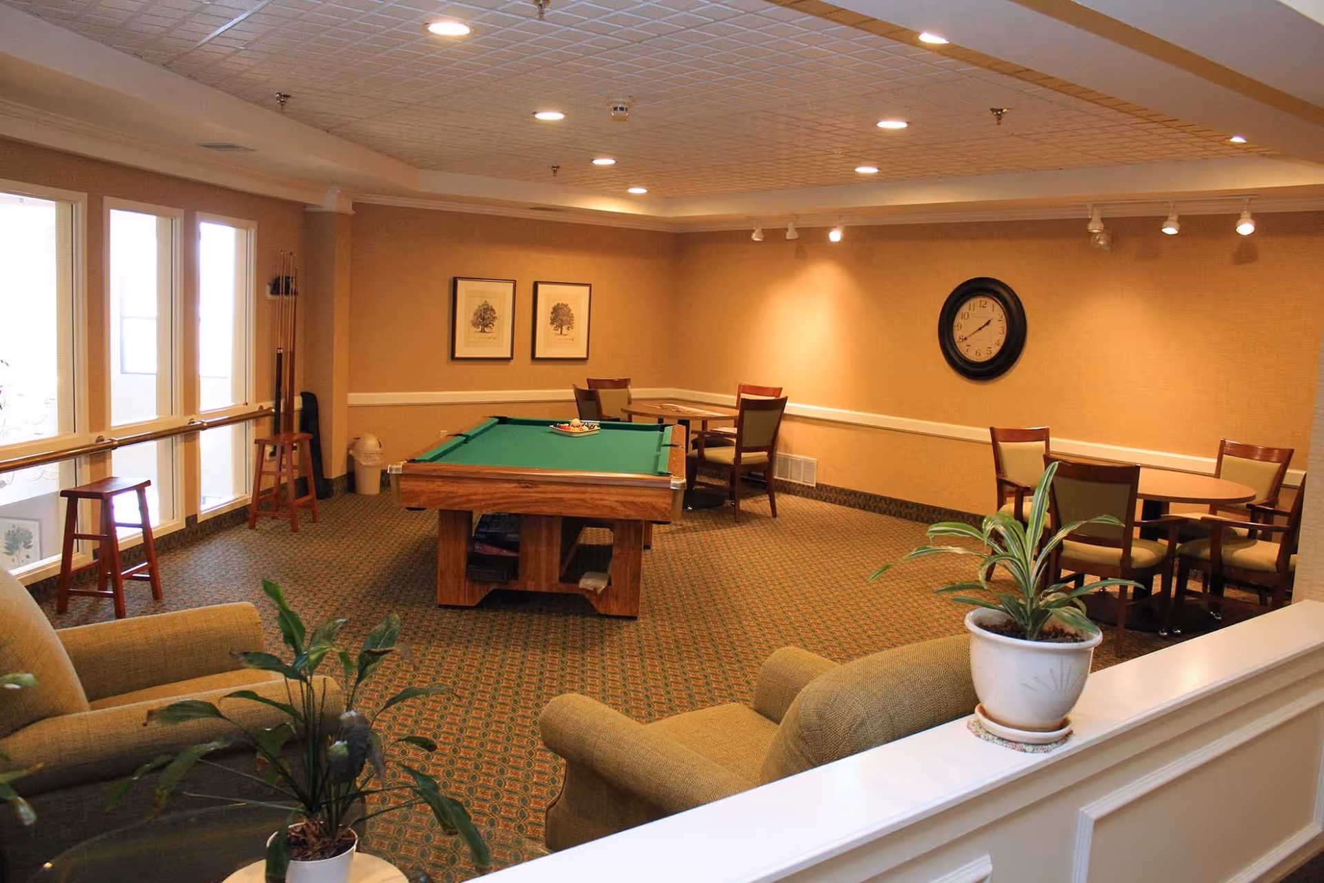 A cozy common room with a pool table in the center, surrounded by chairs and small round tables. The room has beige walls with framed artwork and a clock, carpeted floor, and large windows letting in natural light. There are also two armchairs and potted plants adding to the comfortable atmosphere.