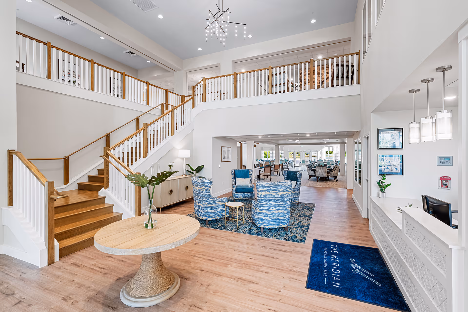 Bright and spacious senior living facility lobby with wooden staircase and white railings, a round wooden table with a vase of green leaves, blue patterned armchairs arranged on a blue rug, and a reception desk with hanging pendant lights. The floor mat near the desk reads 'The Meridian at Punta Gorda Isles.'