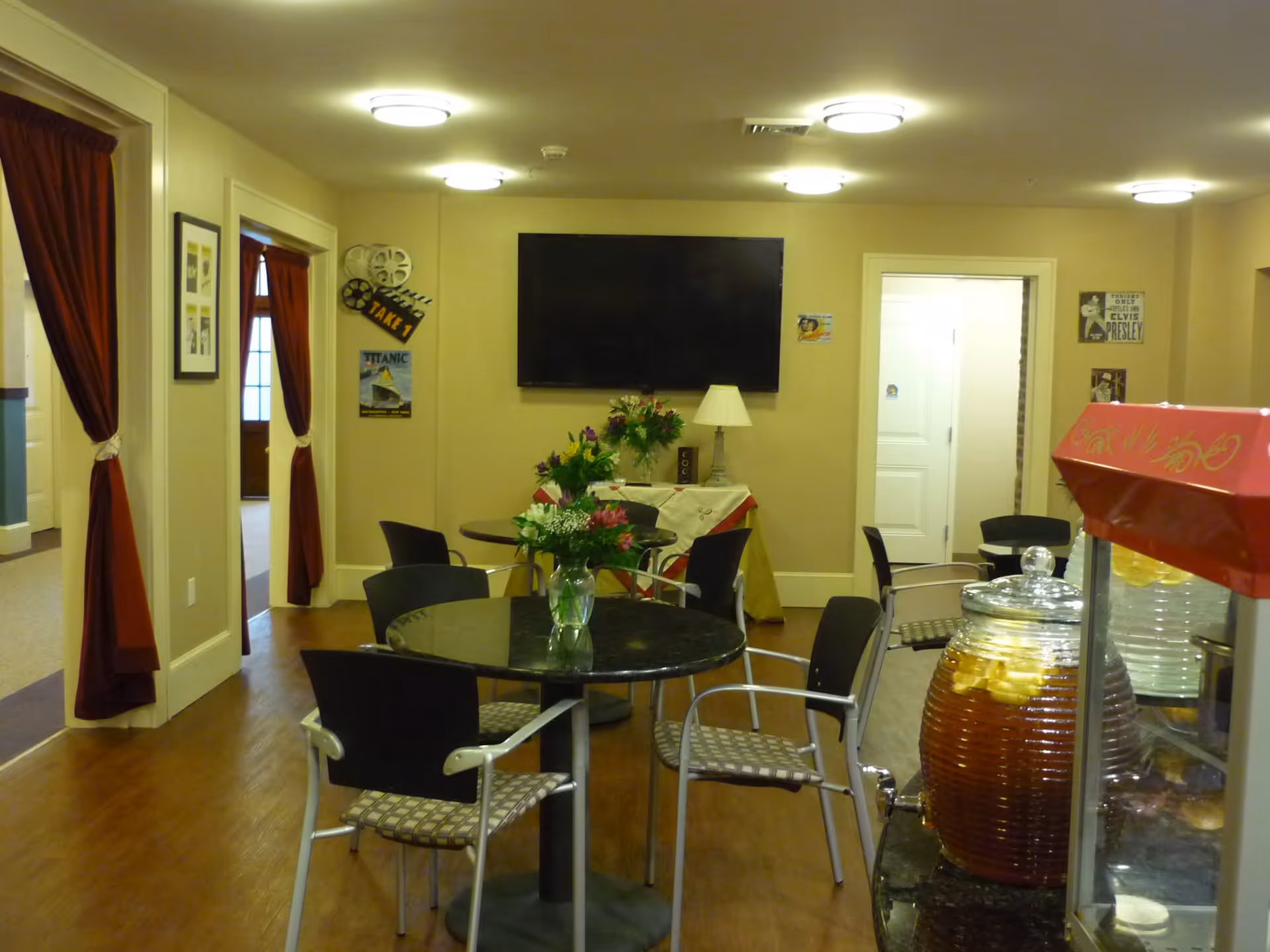 Interior room with round tables and chairs, each table decorated with a vase of flowers. A large flat-screen TV is mounted on the wall, with a lamp and small table underneath. The room has beige walls and wooden flooring. Red curtains frame doorways leading to other rooms. A large glass container filled with iced tea and lemon slices is visible on the right side.