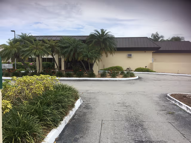 Exterior view of Sunny Hills Assisted Living Facility Homestead showing a single-story building with a dark roof, surrounded by palm trees and landscaped greenery along a paved driveway under a cloudy sky.