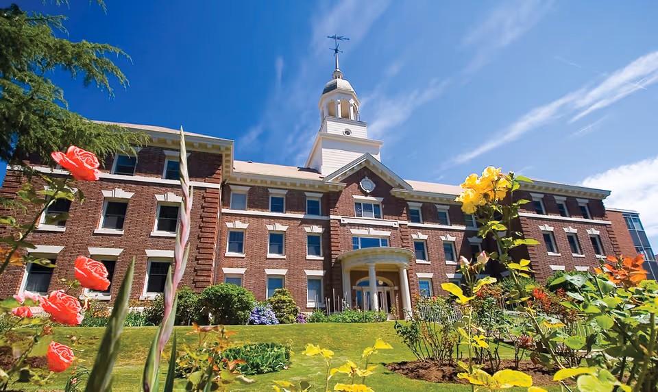 Front exterior view of a large brick building with multiple windows and a central entrance topped by a small tower with a weather vane. The foreground features a garden with colorful flowers and green shrubs under a bright blue sky with wispy clouds.