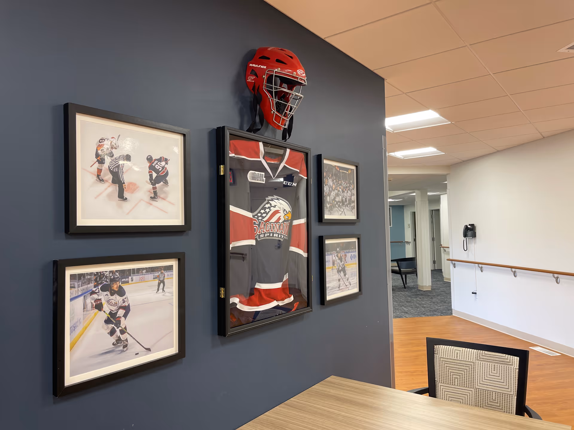 Hallway interior showing a dark blue wall decorated with a framed hockey jersey, helmet, and photos, with a table and chair in the foreground and a corridor to the right.