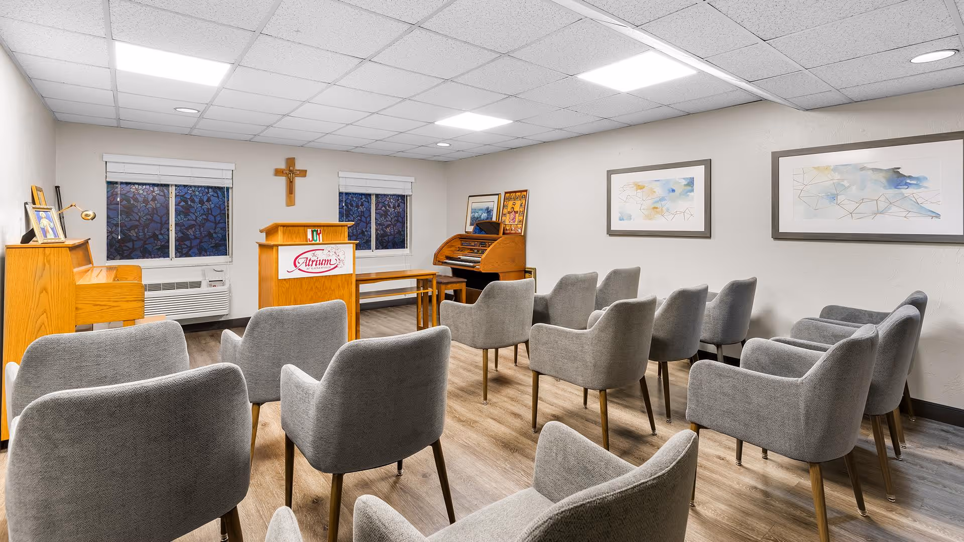 A small chapel or meeting room with rows of gray upholstered chairs facing a wooden podium with the Atrium logo. The room has two windows with blinds, a wooden piano on the left, an organ on the right, a wooden cross on the wall, and two abstract paintings on the right wall. The floor is wood and the ceiling has recessed lighting.