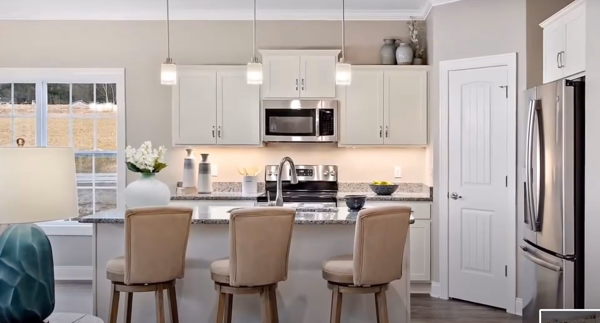 Modern kitchen with white cabinets, granite countertops, and stainless steel appliances including a microwave and refrigerator. Three beige cushioned bar stools are placed at the kitchen island, which has a sink and a vase with white flowers. Pendant lights hang above the island, and a window shows an outdoor view.