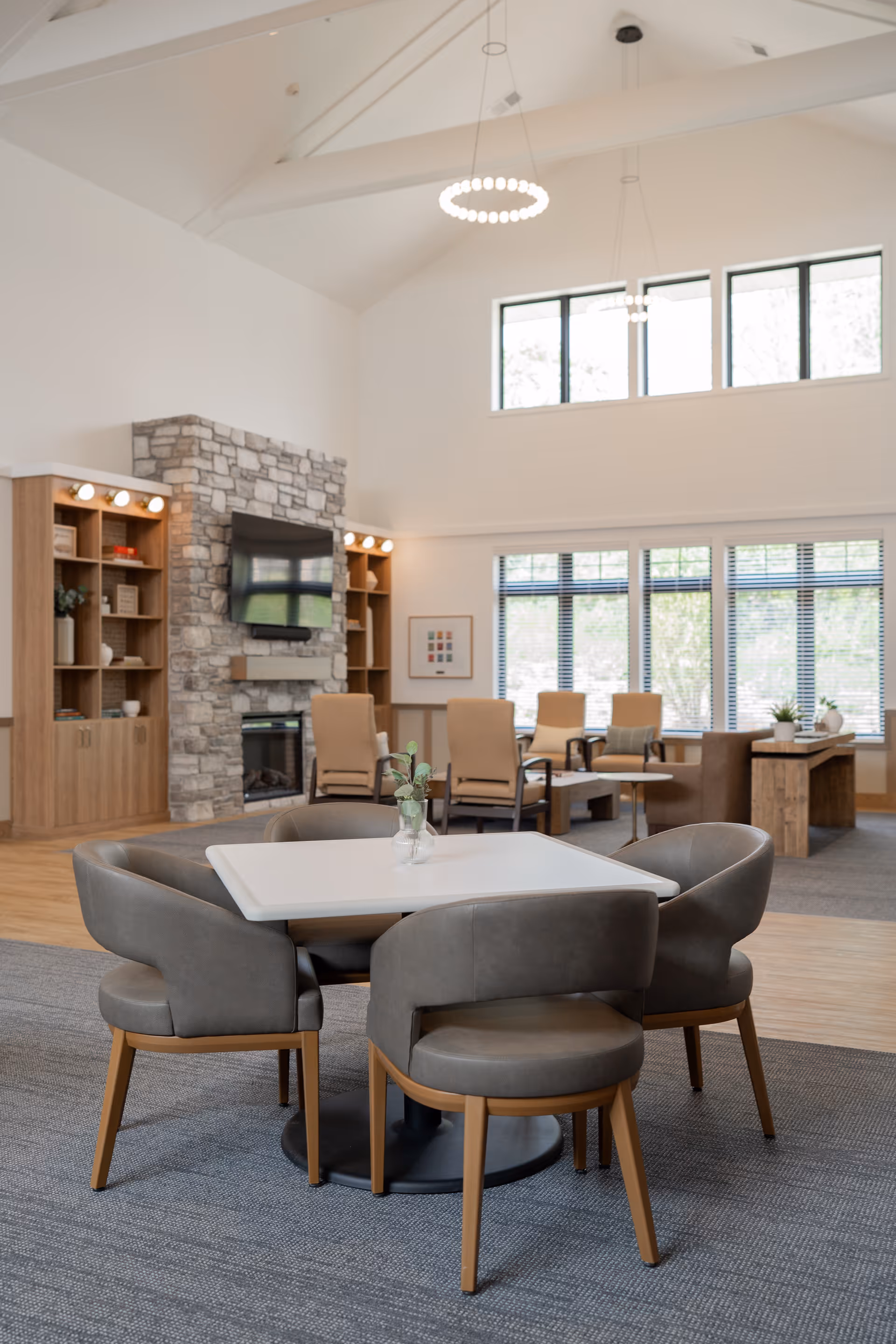 Bright communal living room with a square table and four gray chairs in the foreground, lounge seating, stone fireplace and TV in the background.