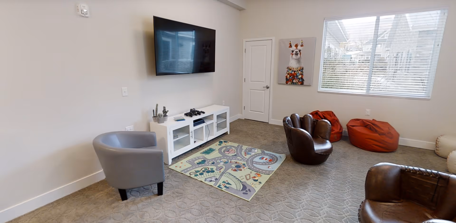 Bright common room with a wall-mounted TV above a white console, several chairs and bean bags, and a play rug beneath a large window.