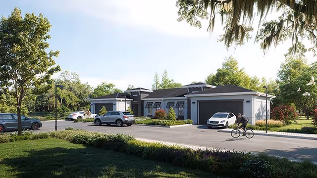 Exterior view of a modern single-story building with a driveway and several parked cars. A person is riding a bicycle on the road in front of the building. The area is surrounded by trees and greenery under a clear sky.