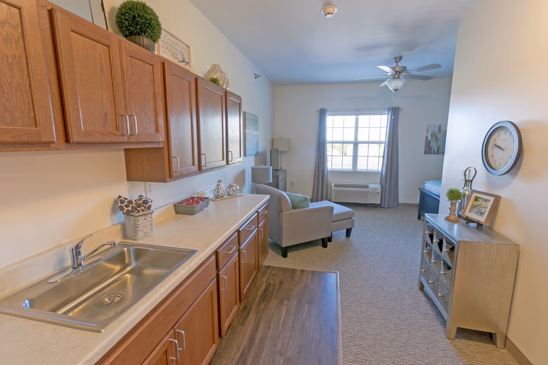 Interior view of a senior living facility room featuring a kitchenette with wooden cabinets and a stainless steel sink on the left. The room extends into a living area with a gray armchair, a window with curtains, a ceiling fan, and a small dresser with decorative items and a wall clock on the right.