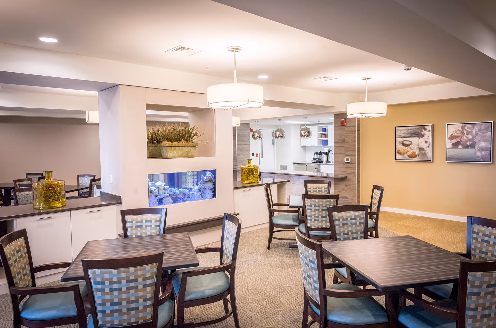 A well-lit dining area in a senior living facility with several square wooden tables surrounded by cushioned chairs featuring a checkered pattern. The room has beige walls adorned with framed artwork and a built-in aquarium with plants above it. Modern pendant lights hang from the ceiling, and a kitchen or service area is visible in the background.