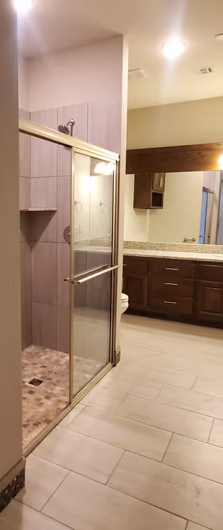 A bathroom with a glass-enclosed tiled shower, light floor tiles, and a granite-top vanity with dark wood cabinets and mirror.