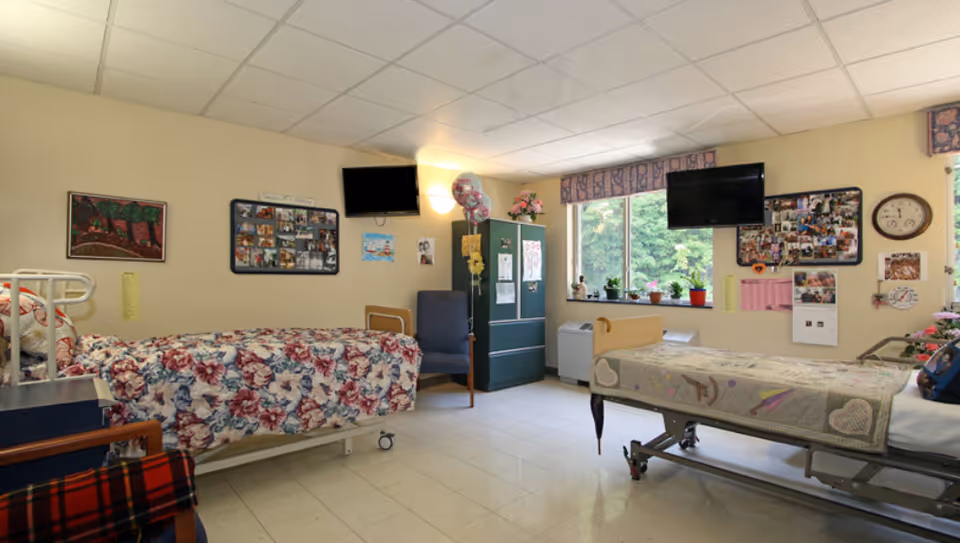 A bright and clean assisted living bedroom with two beds, each with floral and patterned bedspreads. The room has a window with plants on the sill, two wall-mounted televisions, a green cabinet, a blue chair, and various photos and decorations on the walls.