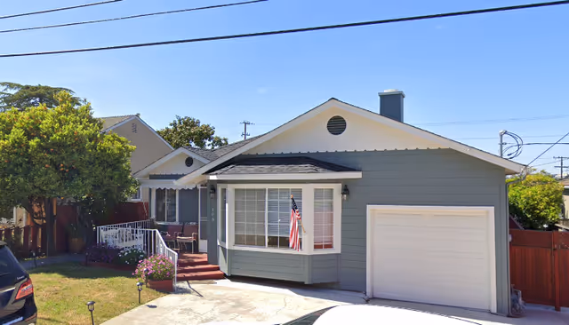 Single-story gray house with an attached garage, front porch displaying an American flag, driveway and small lawn under a clear sky.