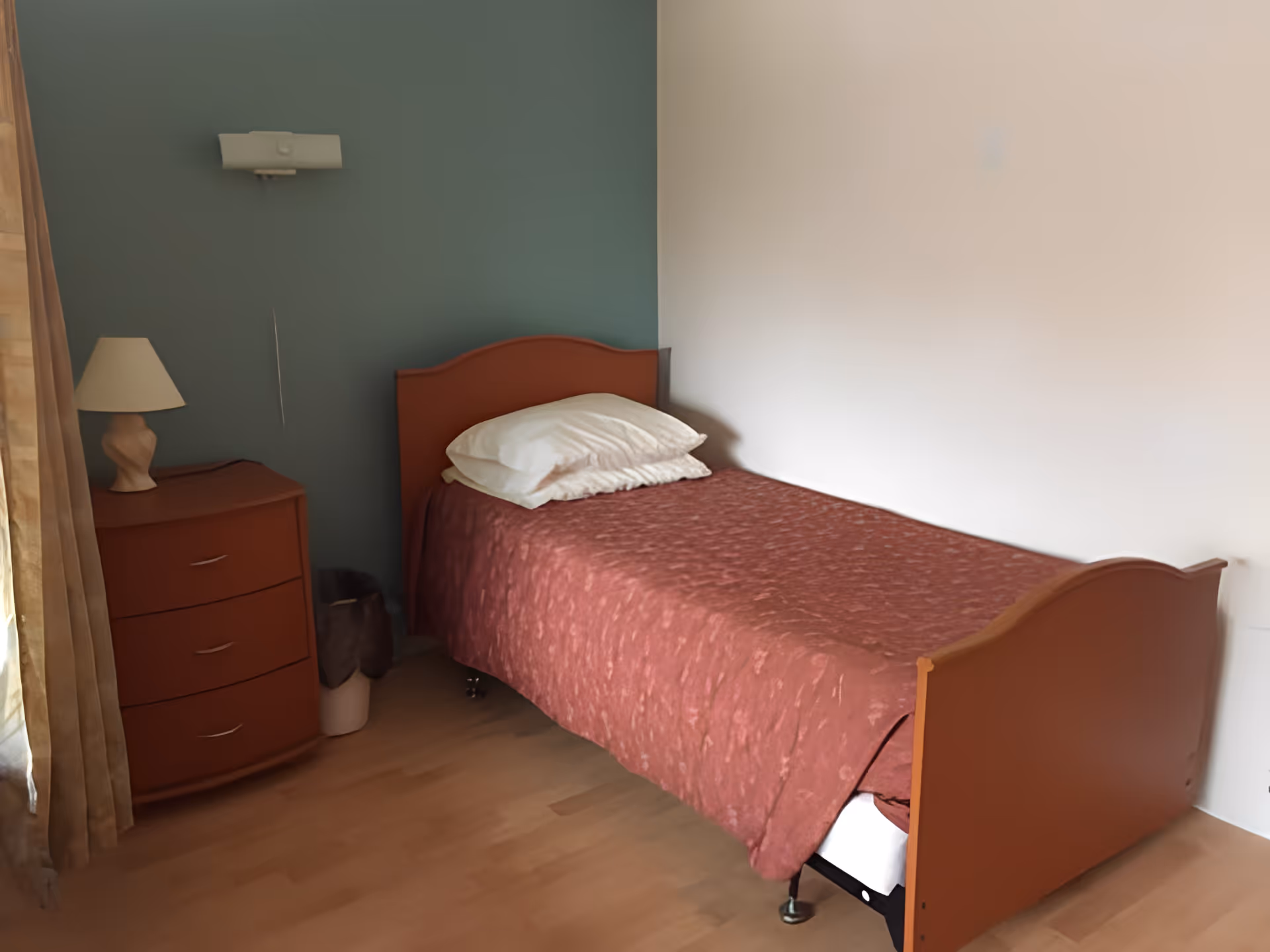 A single bed with a red bedspread and pillows next to a nightstand with a lamp in a simple bedroom.