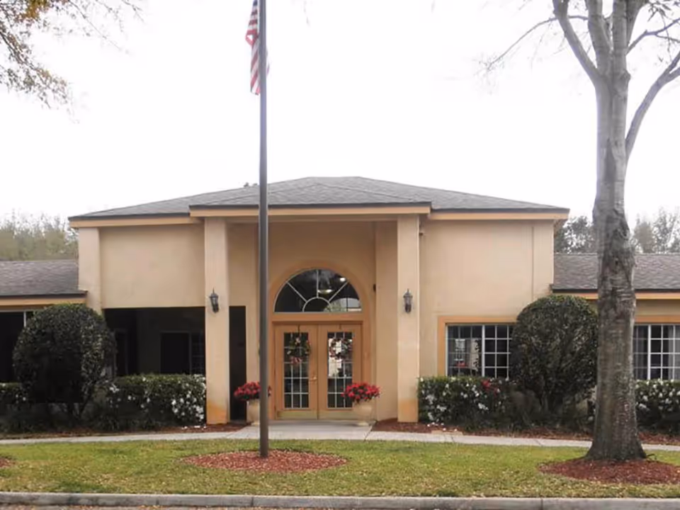 Front exterior view of a single-story building with beige walls and a dark shingled roof. The entrance features double glass doors with an arched window above, flanked by two columns. There are bushes and flowers on either side of the entrance, a flagpole with an American flag in front, and a large tree to the right.