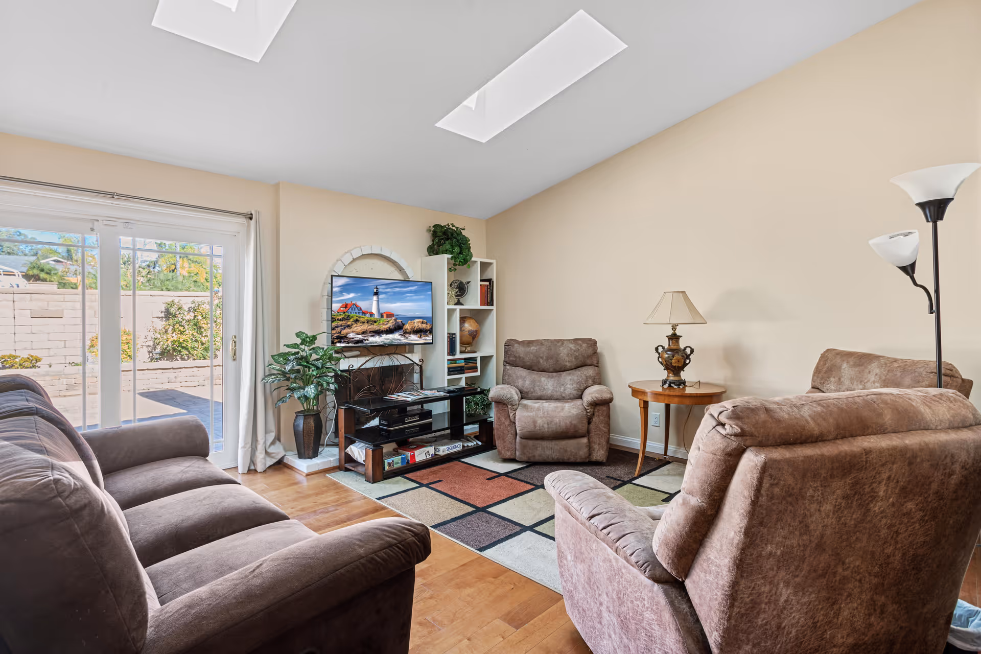 A cozy living room with three brown upholstered armchairs and a matching sofa arranged around a colorful geometric rug. A flat-screen TV is mounted on the wall above a black entertainment unit with shelves holding books and decorative items. There is a wooden side table with a lamp, a tall floor lamp, and a potted plant near sliding glass doors that lead to an outdoor area. The room has light beige walls and skylights on the ceiling.
