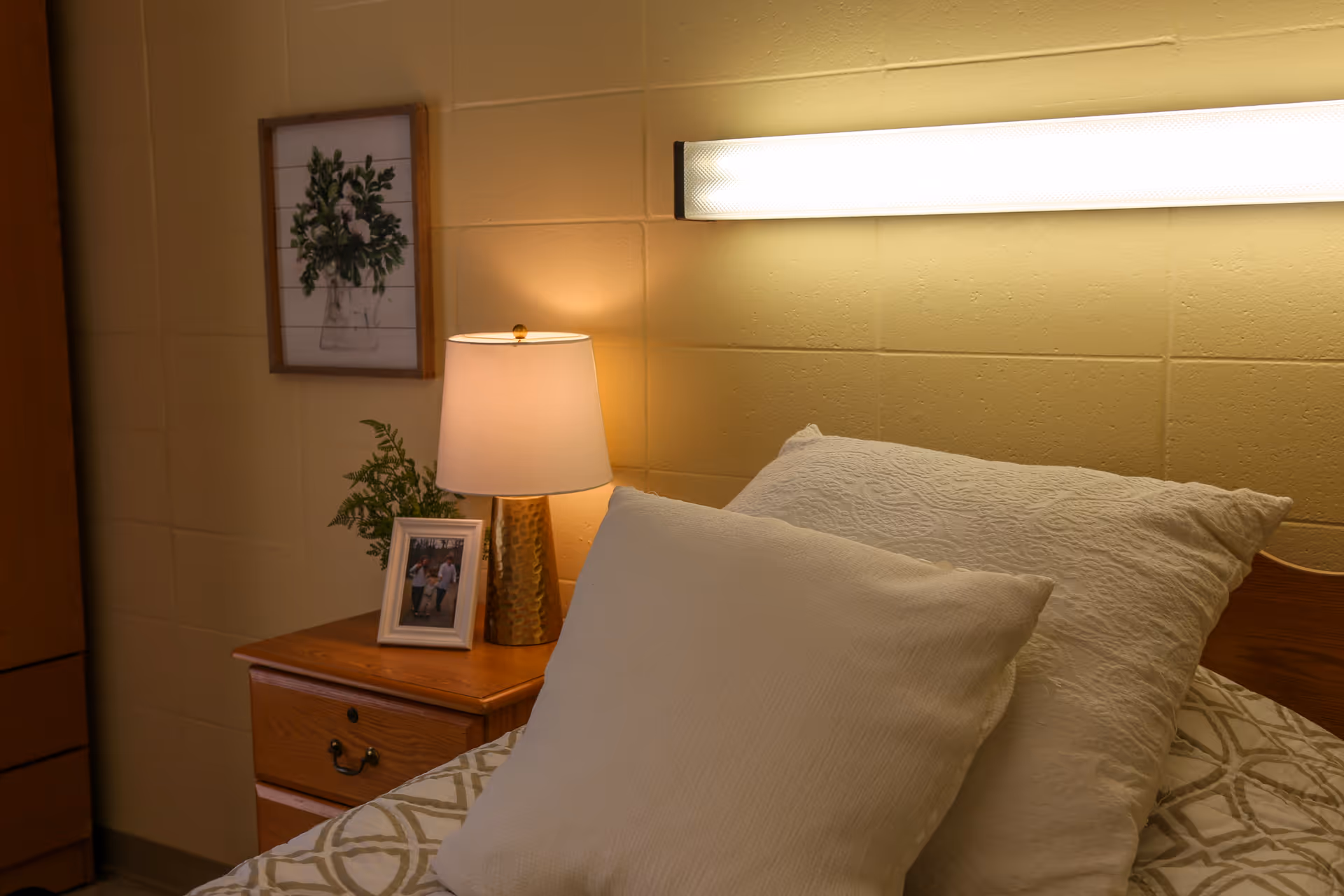 A cozy bedroom corner with a bed featuring white pillows and a patterned bedspread. Next to the bed is a wooden nightstand with a gold lamp, a framed photo, and a small plant. A framed floral artwork hangs on the beige wall above the nightstand, and a long fluorescent light fixture is mounted on the wall above the bed.