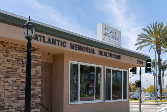 Exterior front view of Atlantic Memorial Healthcare Center building with a stone wall section, large windows, a street lamp, palm trees, and a clear blue sky.