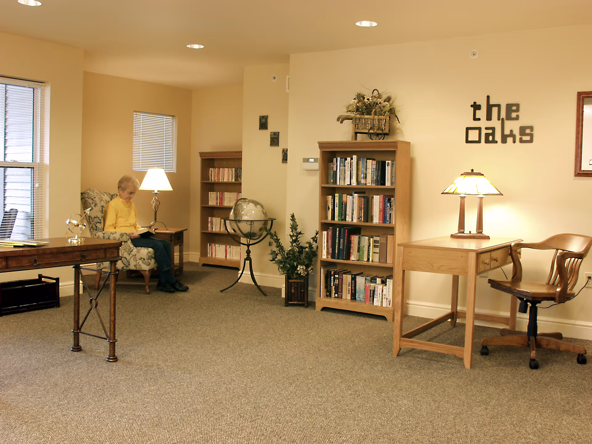 A cozy reading area in The Oaks Retirement Residence featuring a woman sitting in a floral armchair reading a book. The room has beige walls, carpeted floor, two wooden bookshelves filled with books, a globe on a stand, a wooden desk with a lamp and chair, and a wall decoration spelling 'the oaks'.