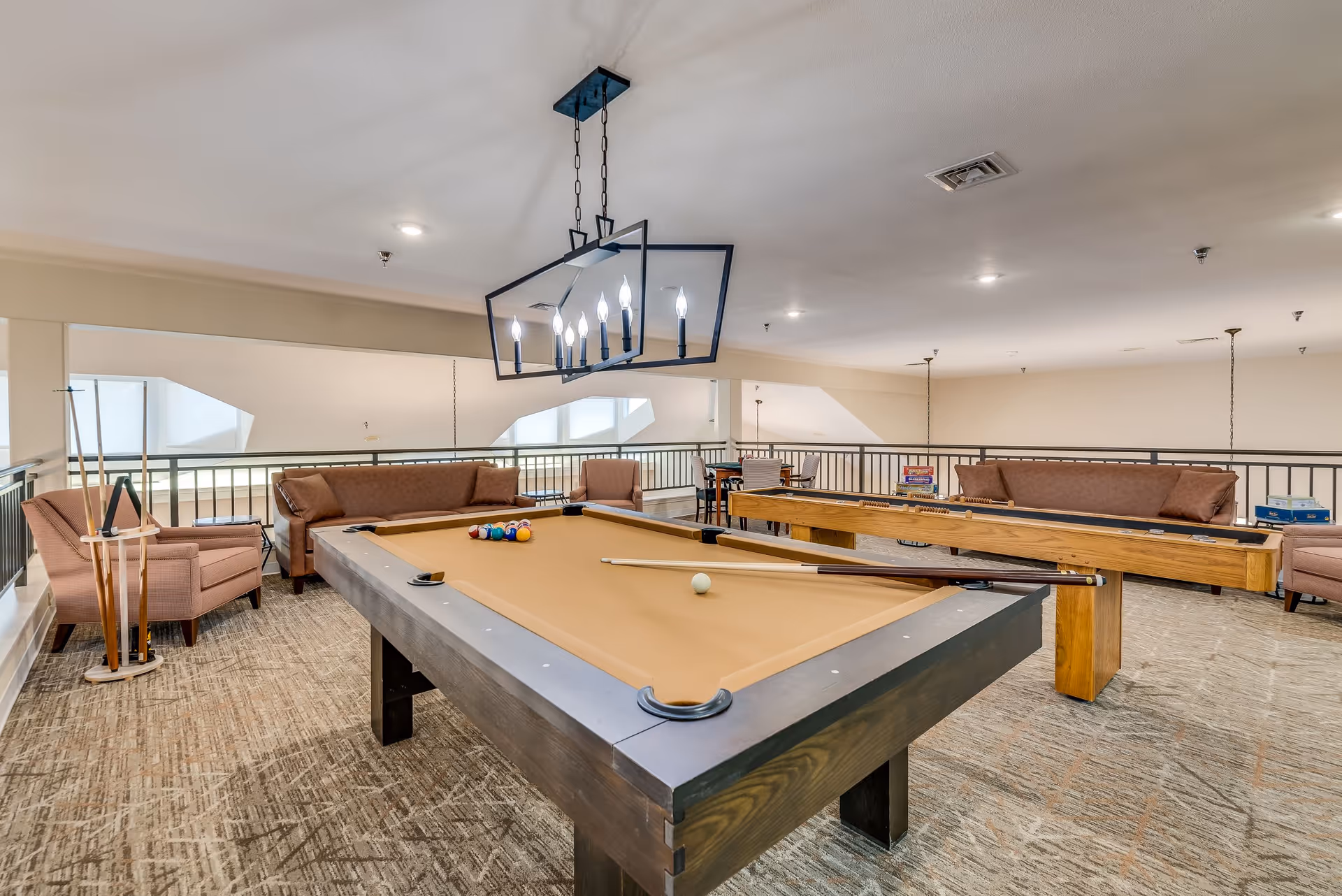 A bright interior recreation room featuring a pool table, shuffleboard table, sofas and chairs under a modern chandelier.