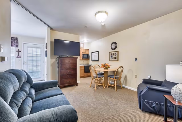Interior view of a senior living facility room featuring a blue upholstered sofa on the left, a dark armchair on the right, a wooden dining table with four chairs in the center, and a flat-screen TV mounted on a tall wooden dresser. The room has beige walls and carpeted flooring, with a small kitchen area visible in the background.