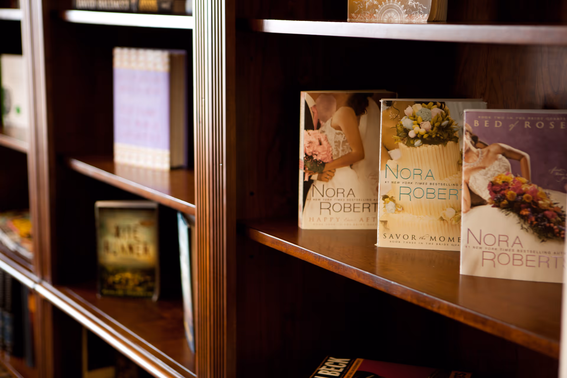 Close-up view of a wooden bookshelf displaying several books, including multiple novels by Nora Roberts with covers featuring wedding and floral themes.