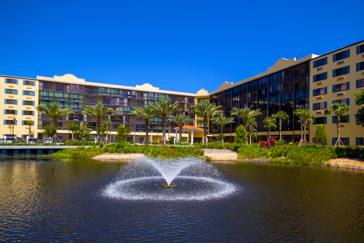 View of a large senior living facility building named Village On The Isle with multiple floors, large windows, and palm trees in front. There is a pond with a water fountain in the foreground under a clear blue sky.