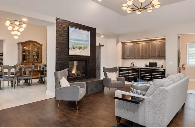 A cozy living room area with a gray sofa, two gray armchairs, and a modern fireplace with a painting above it. To the left, there is a dining area with a wooden dining table and chairs, and a glass-fronted cabinet. The room has wooden flooring and light-colored walls, with a small kitchenette area featuring cabinets and a countertop in the background.