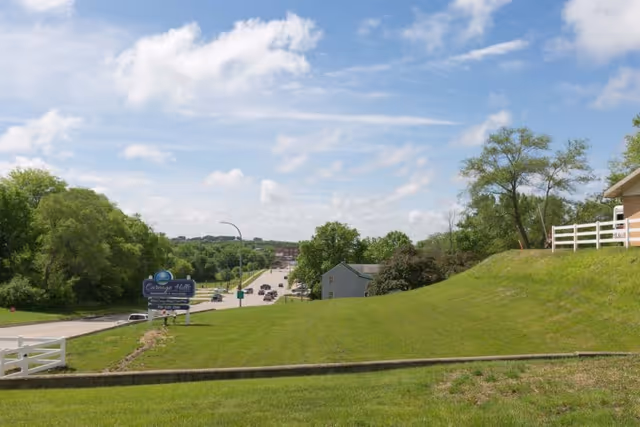 A grassy hill with a white fence on the right side and trees in the background. A road with several cars is visible in the distance, along with a sign for Careage Hills Rehabilitation And Healthcare on the left side near the road. The sky is partly cloudy with blue patches.