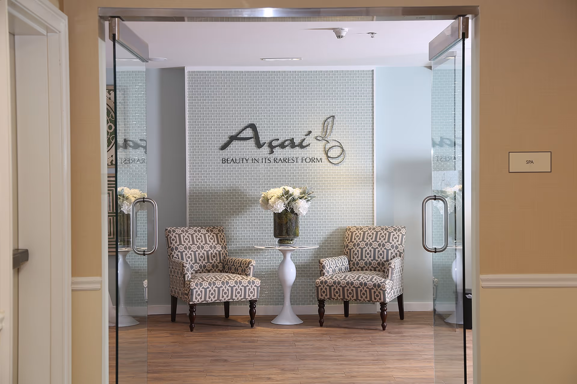 A small seating area inside a spa with two patterned armchairs and a white round table between them holding a vase of white flowers. The wall behind has a sign that reads 'Açaí BEAUTY IN ITS RAREST FORM'. The entrance has glass doors and a sign on the right wall labeled 'SPA'.