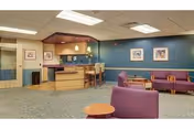 A senior living facility lounge with purple armchairs and small tables facing a reception desk beneath teal walls and framed artwork.
