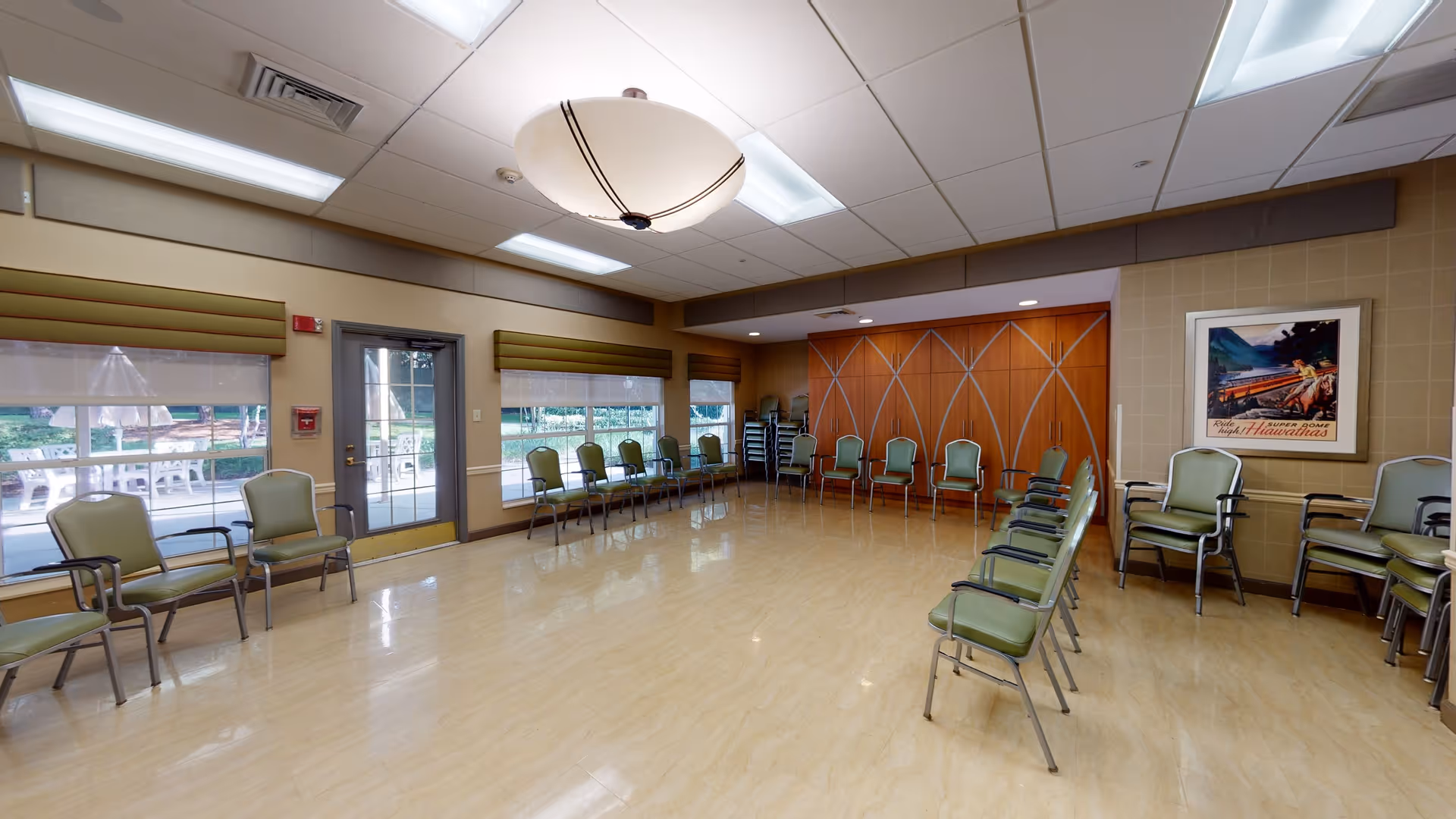 A spacious room with a polished floor and green cushioned chairs arranged along the walls. Large windows with green valances let in natural light, and a glass door leads outside to a patio area with tables and chairs. The ceiling has recessed lighting and a large central light fixture. A framed vintage-style poster hangs on one wall.