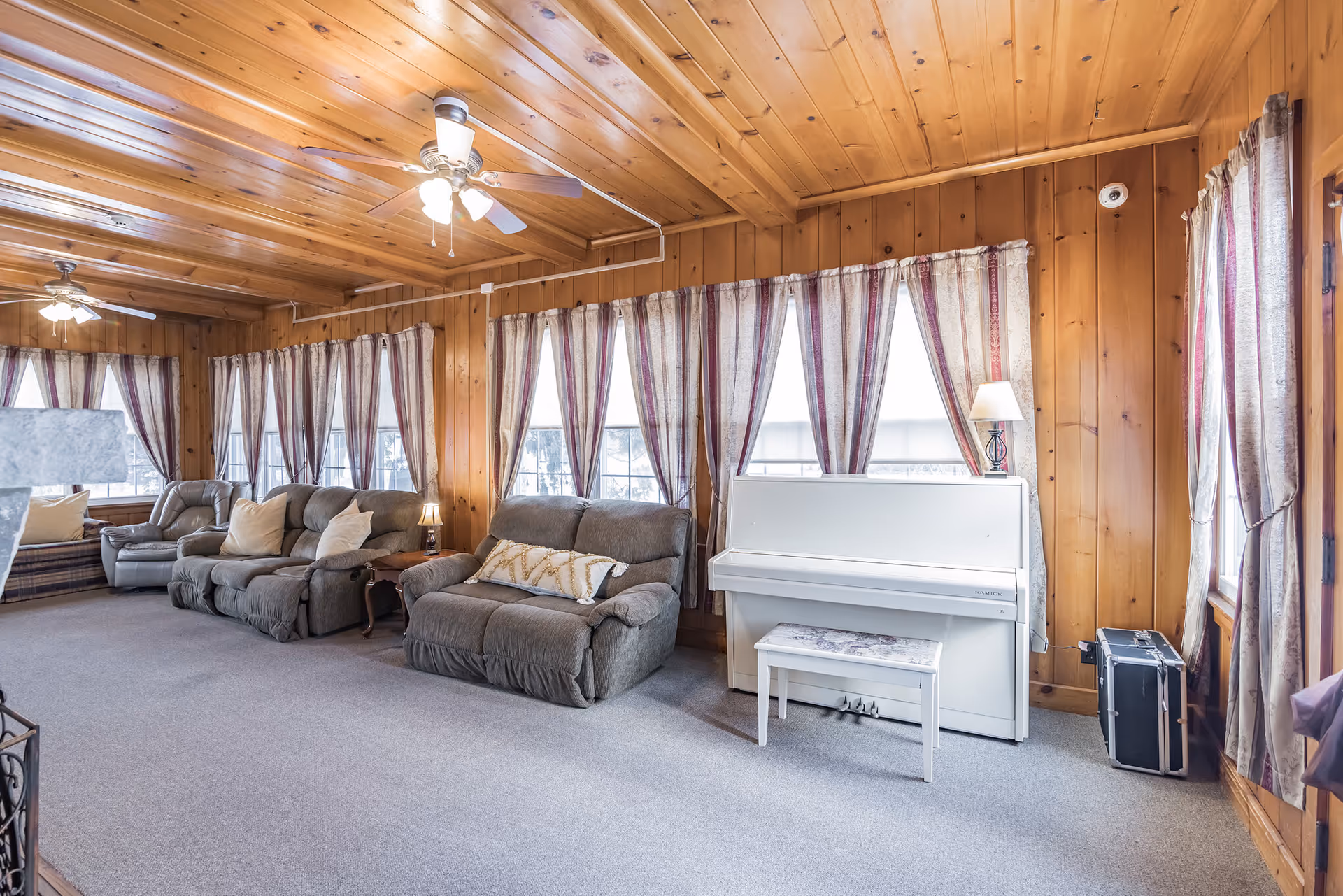 A cozy living room with wood-paneled walls and ceiling, featuring multiple windows with striped curtains. The room contains several cushioned recliner chairs and sofas, a white upright piano with a matching bench, a small lamp on the piano, and a ceiling fan with lights.