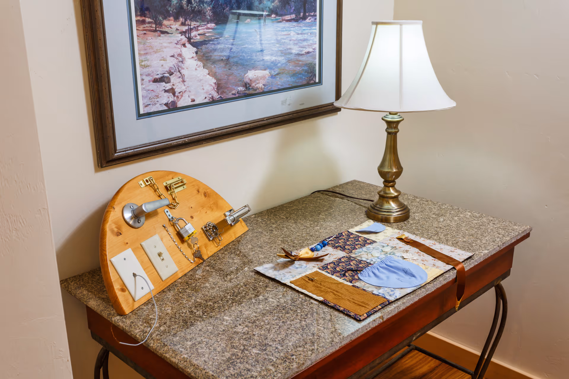 Side table topped with a brass lamp, a wooden activity board with locks and switches, and a fabric placemat beneath a framed landscape print.