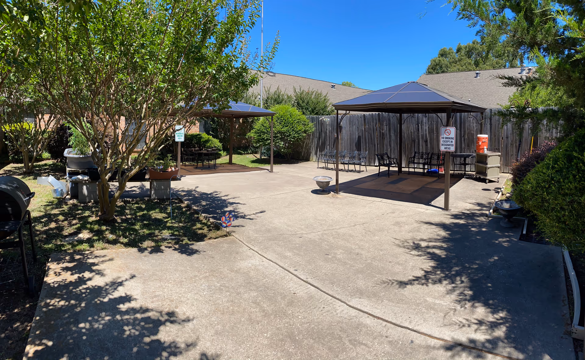 Concrete courtyard with two covered pavilions, benches, trees and a wooden fence under a clear blue sky.
