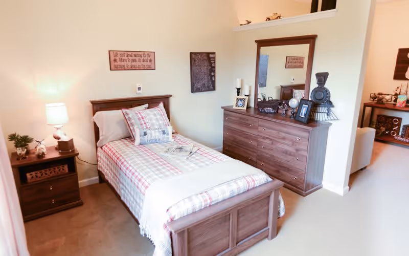 A cozy bedroom with a single bed featuring plaid bedding and a decorative pillow. Next to the bed is a wooden nightstand with a lamp, a small plant, and decorative items. Across from the bed is a wooden dresser with a large mirror, candles, framed photos, and decorative objects. The room has beige walls and carpeted flooring, with a partial view into an adjacent living area.