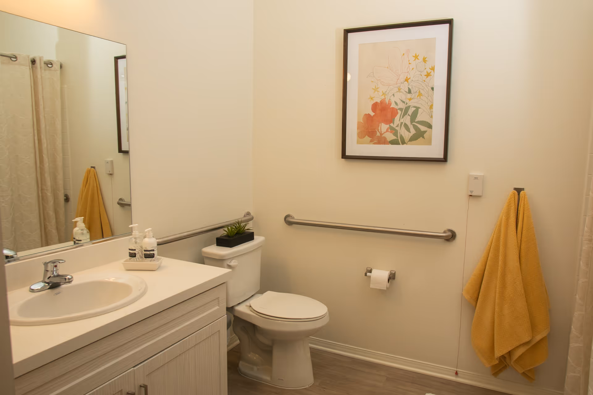 A clean bathroom with a white sink and countertop, a toilet with a small plant on the tank, a large mirror above the sink, a framed floral artwork on the wall, yellow towels hanging on a hook, and a grab bar installed on the wall next to the toilet.