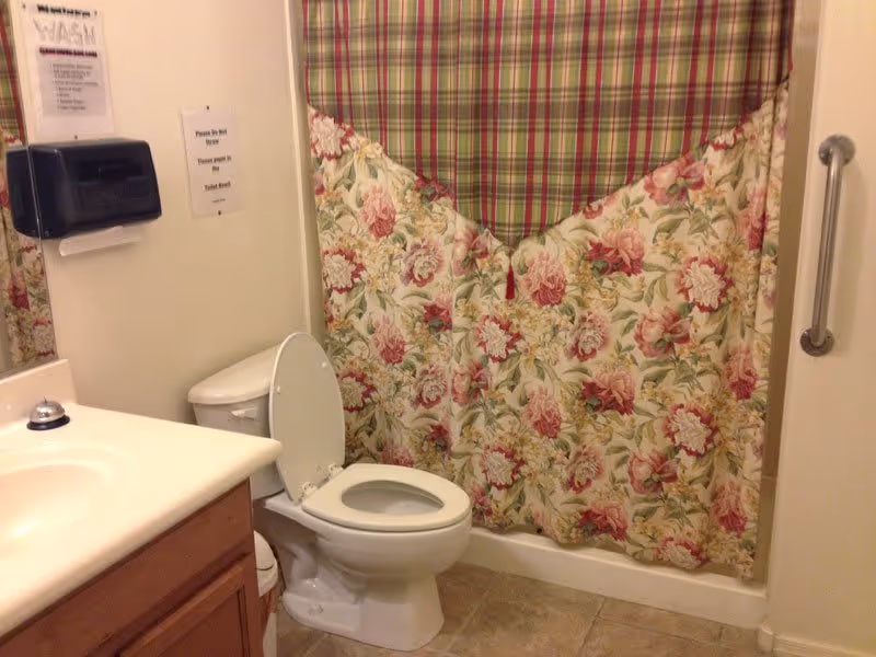 A bathroom with a white toilet with the lid open, a sink with a countertop and wooden cabinet, a floral and plaid shower curtain, a grab bar on the wall, and a paper towel dispenser mounted on the wall.