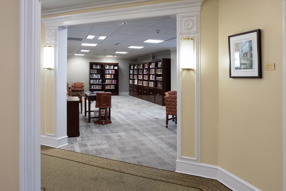 View through a doorway into a quiet library room with bookshelves filled with books, several wooden tables and chairs, and upholstered armchairs. The walls are light-colored with framed artwork and wall sconces providing soft lighting.
