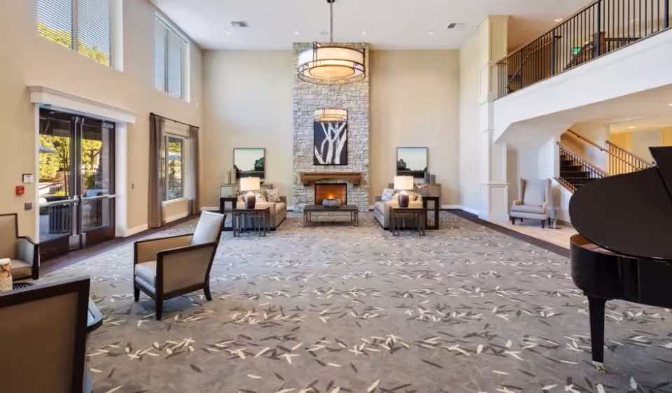 Spacious living room area with high ceilings, large windows, and a stone fireplace in the center. The room features two sofas with side tables and lamps, several armchairs, a grand piano on the right, and a staircase leading to an upper level. The room is well-lit with natural light and modern hanging light fixtures.