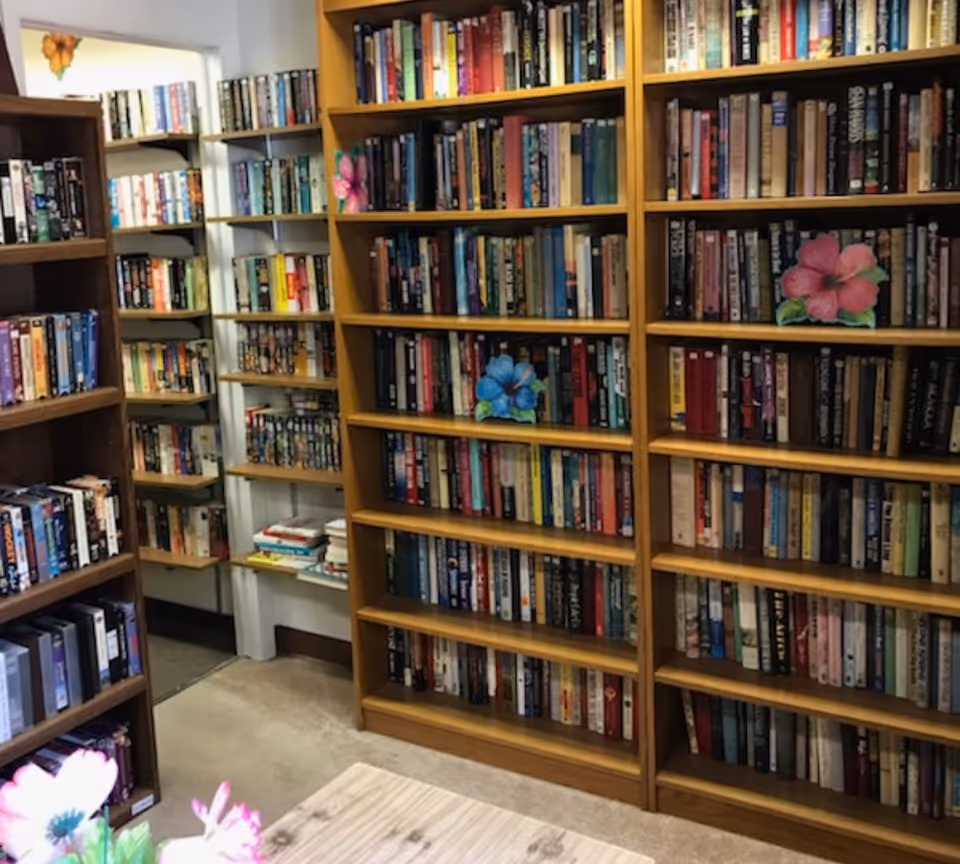 Interior view of a small library or reading room with wooden bookshelves filled with books. There are colorful flower decorations placed on some shelves and a wooden table in the foreground.