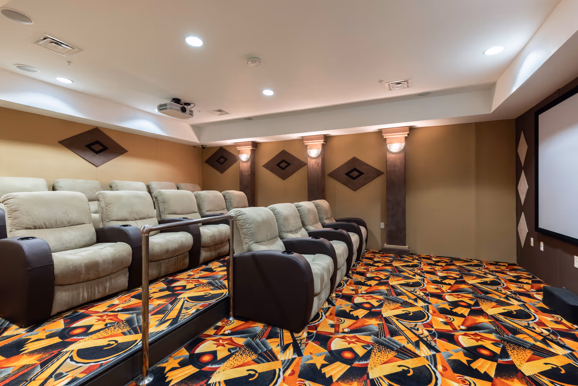 A cozy senior living facility theater room with two rows of plush beige recliner chairs with dark armrests, a colorful patterned carpet, wall sconces, and a large projector screen on the right wall.