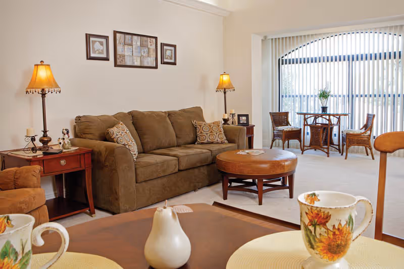 A cozy living room with a brown sofa adorned with patterned cushions, two side tables each with a lamp, a round leather ottoman, and a small dining area with a round glass table and wicker chairs near a large window with vertical blinds.