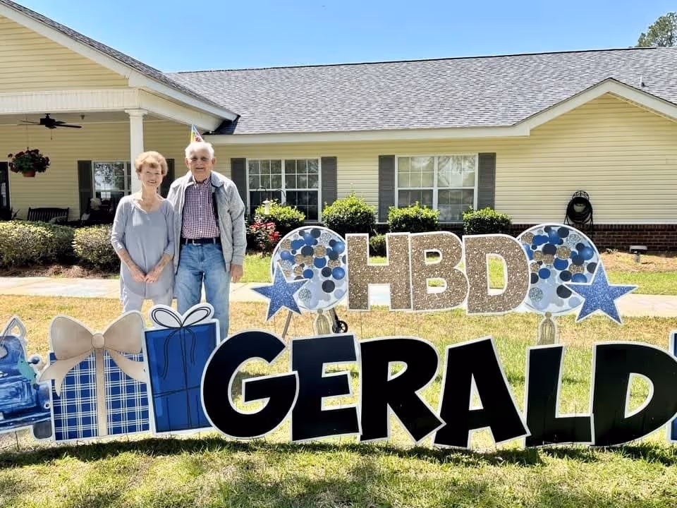 An elderly couple standing outside in front of a single-story yellow building with a porch. In front of them is a large decorative sign on the grass that reads 'HBD GERALD' with birthday-themed decorations including stars, balloons, and gift boxes.