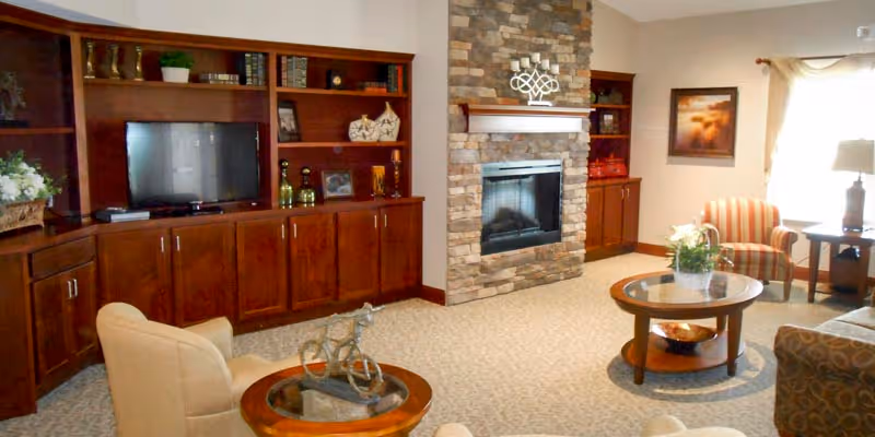 A cozy living room with beige carpet, a stone fireplace, wooden built-in shelves with a TV, and comfortable seating including an armchair and a striped chair near a window with curtains. There are two round wooden coffee tables with decorative items on them.