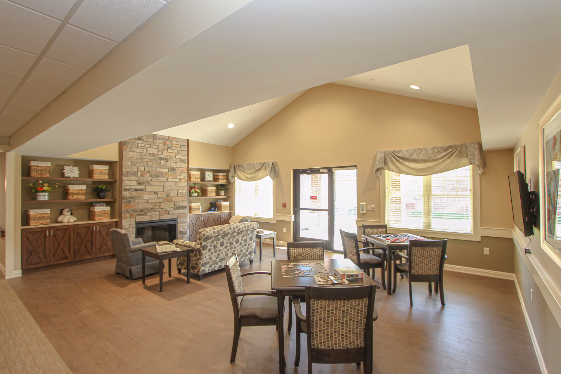 A bright and spacious common area in a senior living facility featuring a stone fireplace, built-in shelves with decorative baskets and plants, multiple tables with chairs arranged for games or puzzles, and large windows with valances allowing natural light to fill the room.
